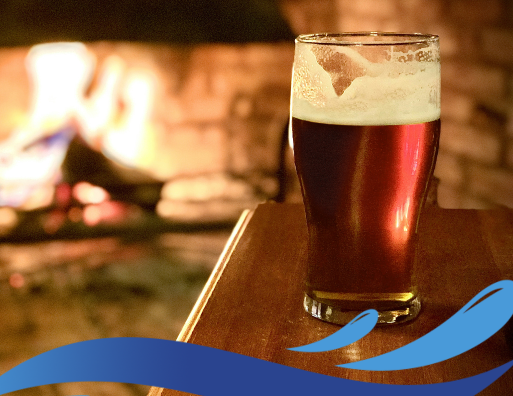 Glass of beer on a table in front of a fireplace. Photo by Dan Barrett at Unsplash
